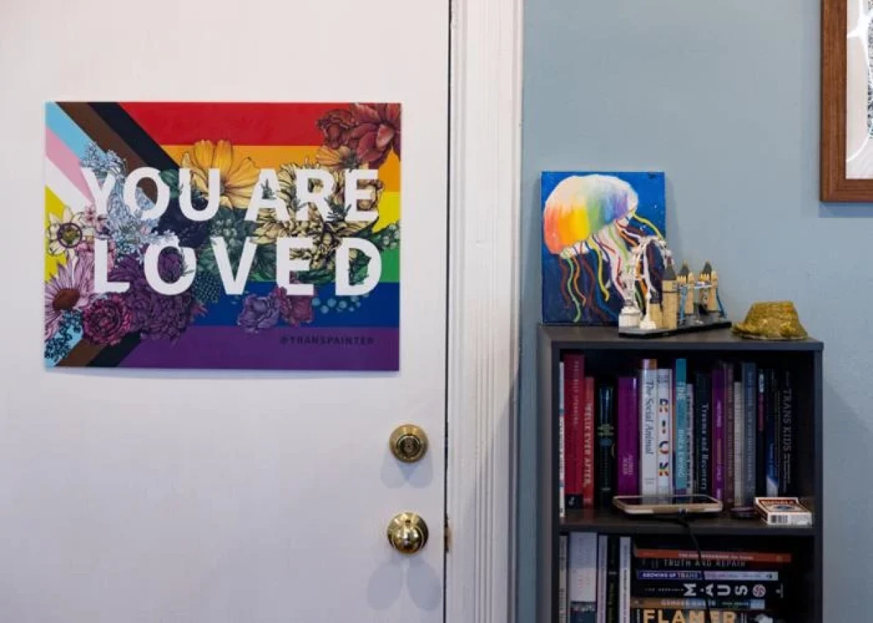 A progress pride flag with the words 'you are loved' hangs on the back of a door. There's a bookcase full of books next to the doorframe. On top is a model castle and a painting of rainbow jellyfish behind it. 